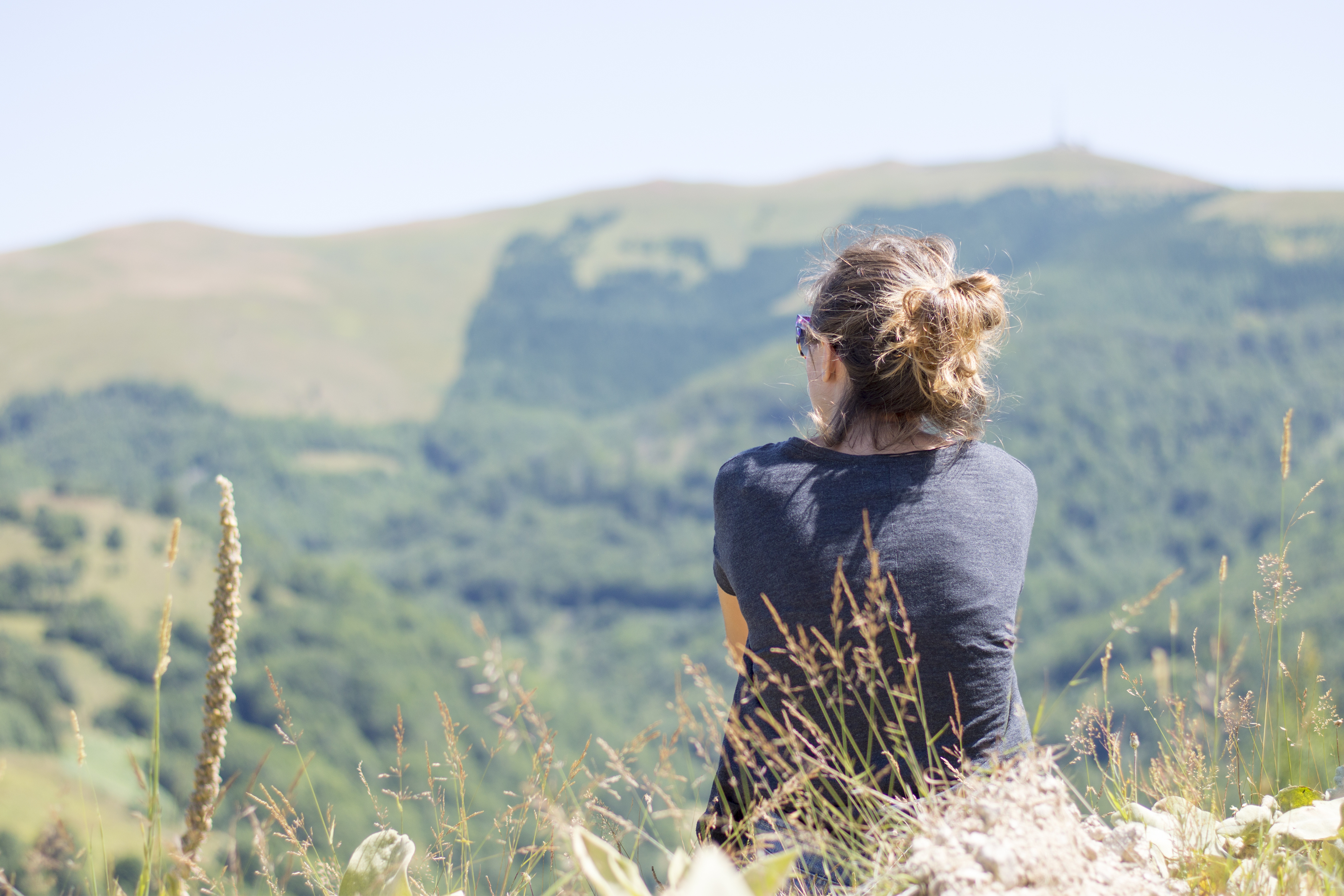 Young woman with backpack sitting on cliff's edge and looking to mountains and clear sky. Summer hiking trip adventure
